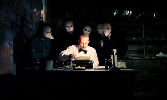 A picture of Sleep No More - an actor sits at a desk in shirtsleeves and a bowtie, examining a sheet of paper sitting in a typewriter. A desklamp illuminates his face. Behind him, in darkness, several masked figures crowd, peering at him, or over his shoulder - the audience of this interactive play.